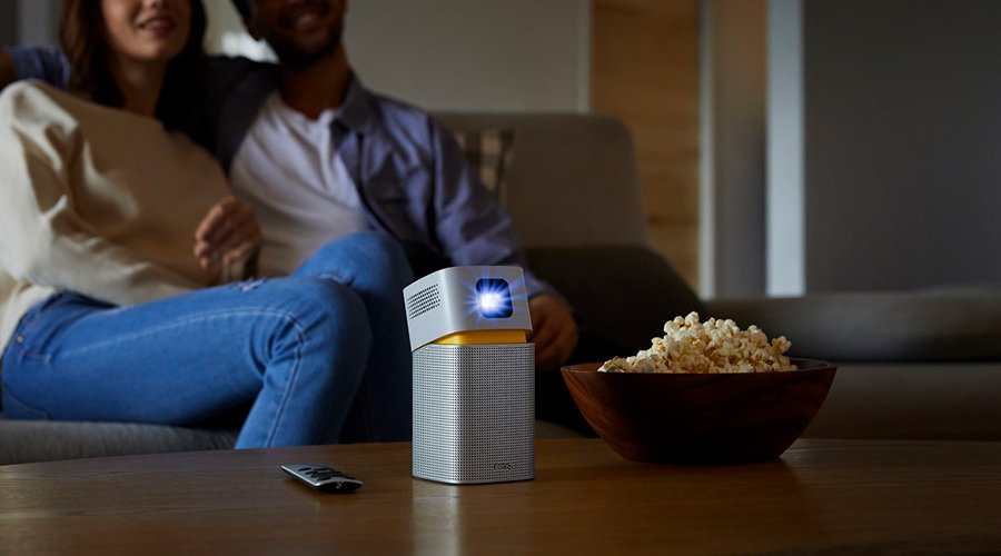 A portable projector on the table with a couple sitting on the couch in the background
